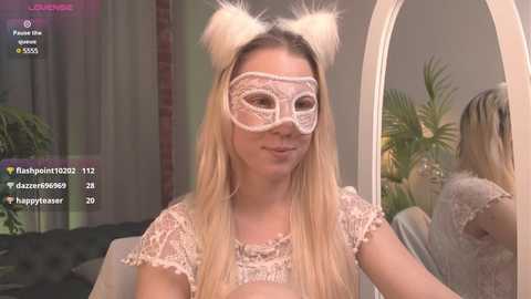 Media: Video of a blonde woman in a white lace mask and cat ears, wearing a delicate lace top, seated in a modern living room with a mirror and greenery.