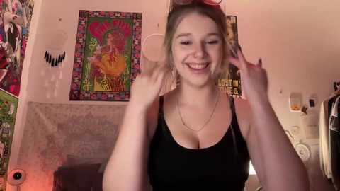 Media: Video of a smiling young woman with fair skin and blonde hair, wearing a black tank top, adjusting sunglasses. Background features colorful tapestries, posters, and eclectic decor.