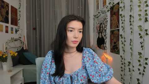 Media: Video of a young woman with long black hair, fair skin, and wearing a blue floral dress, sitting indoors surrounded by decorative items, including flowers and posters.