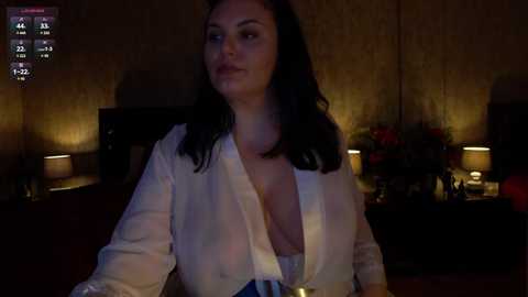 Media: A video of a dimly lit room with a woman in a white blouse, revealing cleavage, sitting at a table. Background shows wooden paneling, a TV, and flowers.