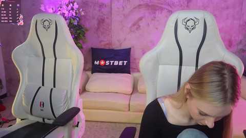 Video of a young woman in a black top sitting on a white gaming chair with a skull logo, surrounded by a beige couch with a pillow saying \"MISTREET,\" in a dimly lit room with purple lighting.