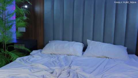 Media: A video of a well-lit bedroom with a neatly made bed featuring two white pillows and a white duvet, set against a dark wood-paneled headboard and a large potted fern on the left.