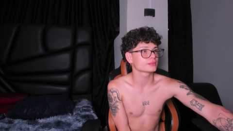 Media: Video of a shirtless, curly-haired young man with tattoos and glasses, sitting on a chair in a dimly lit room with black curtains and a bed with a blue blanket.