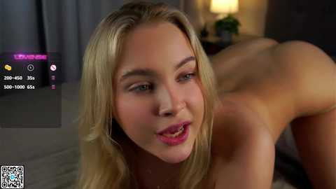 Media: A video of a nude, light-skinned, blonde woman with blue eyes and pink lipstick, bending over on a bed, in a dimly lit room.