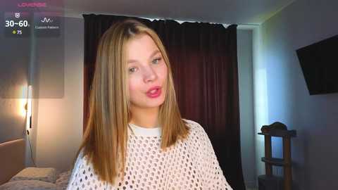 Media: Video of a young Caucasian woman with straight blonde hair, wearing a white crocheted top, standing in a dimly lit bedroom with a black curtain and a small cat on a shelf.