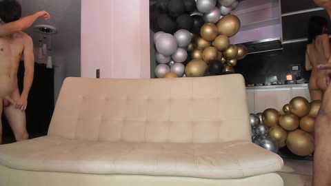 Media: Video of a modern living room with a beige sectional sofa, two nude men, and a pile of metallic balloons in the background.