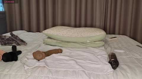 Media: A video of a messy, unmade bed with a cream comforter, a green pillow, a wooden dildo, a black vibrator, and a glass bottle, set against brown curtains in a dimly lit room.