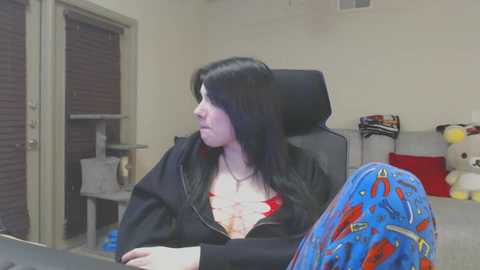 Media: Video of a young woman with long black hair, wearing a black hoodie, sitting at a desk in a cluttered room with toys and a closed door.