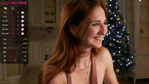 Video of a smiling woman with light skin, auburn hair, and a pink top, standing in a warmly lit room with a decorated Christmas tree in the background.