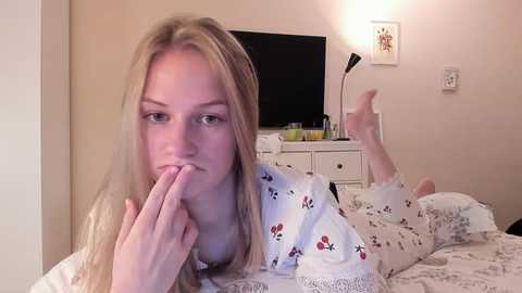 Media: A video of a young blonde woman in a white floral pajama top, lying on a bed, with a finger to her lips, in a softly lit, beige room with a flat-screen TV and a desk lamp.