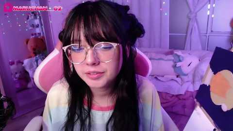 Media: Video of a young woman with long black hair, wearing glasses, in a colorful, cozy bedroom with plush toys, pastel hues, and fairy lights.