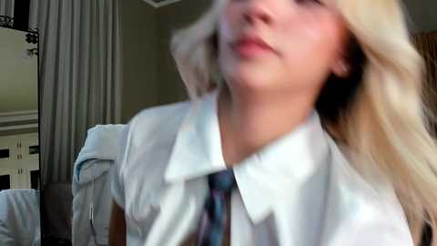 A blurred video of a young, blonde woman in a white blouse with a blue tie, standing indoors with a gray curtain and a white shelf in the background.