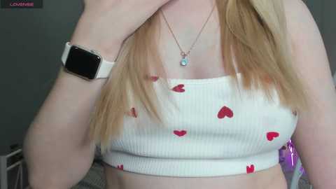 Media: Video of a young woman with fair skin, wearing a white ribbed crop top with red heart patterns and a smartwatch on her left wrist.