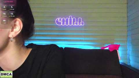 Media: Video of a woman with dark hair tied back, wearing a black shirt, seated on a couch with a red pillow and a glowing \"Sick\" sign in the background.