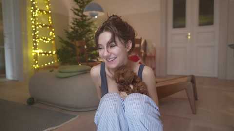 Media: Video of a smiling, dark-haired woman in striped pajamas holding a brown and white dog in a cozy, lit living room with a decorated Christmas tree and fairy lights.