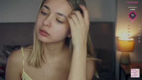 Media: Video of a young Caucasian woman with blonde hair, looking tired, in a bedroom with a lit lamp.