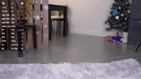 Video of a modern living room with a gray carpet and a fluffy white rug, featuring a small Christmas tree decorated with blue and silver ornaments, and a black chair. A cat is curled up under the tree.