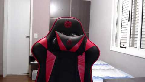 Media: Video of a red and black gaming chair with a large logo, placed in a modern bedroom with white walls, a bed, and a window with white blinds.