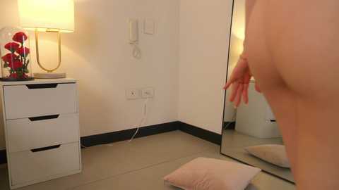 Media: Video of a bare-chested woman with fair skin, medium-sized breasts, and long pink nails, standing in a modern, minimalist room with beige walls, a white dresser, and a large mirror reflecting her reflection.