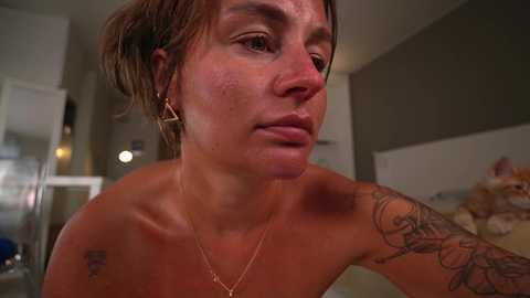 Media: Video of a topless woman with short brown hair, freckles, and a large tattoo on her right shoulder. She has a serious expression, wearing a gold necklace. The background shows a modern, dimly lit kitchen.