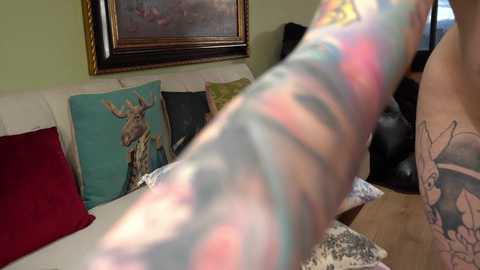 Media: A video of a tattooed arm holding a framed painting, featuring a deer, against a green wall. The arm is positioned prominently in the foreground, showcasing colorful and intricate tattoos.