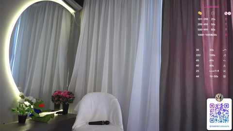 Media: A video of a serene, modern hotel room featuring a white chair draped with a white cloth, a round mirror with a glowing light, and a floral arrangement on a side table.