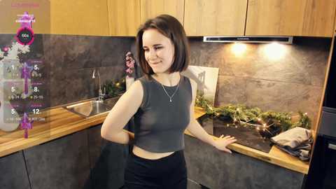 Media: Video of a young woman with straight, shoulder-length brown hair, wearing a sleeveless gray crop top and black pants, standing in a modern kitchen with wooden cabinets, stainless steel appliances, and festive greenery.