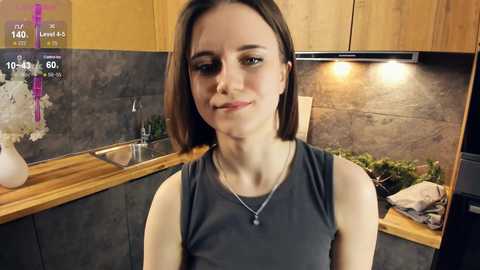 Media: Video of a young Caucasian woman with straight brown hair, wearing a gray sleeveless top, standing in a modern kitchen with wooden cabinets, a backsplash, and potted plants.