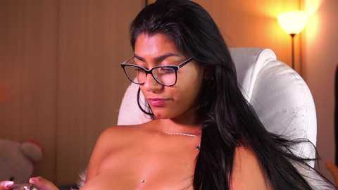 Media: Video of a topless, light-skinned woman with long black hair, wearing black-framed glasses, seated on a white chair in a dimly lit room with a warm, amber light source.