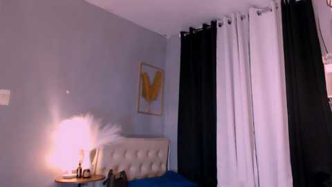 Media: Video of a modern bedroom with a light gray wall, a tufted beige headboard, a lamp with a pink shade, black and white curtains, and a framed abstract art piece.