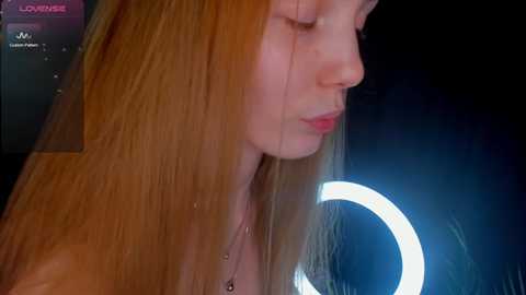 Media: Video of a young woman with long, straight, reddish-brown hair, wearing a black top, standing in a dimly lit room with a bright, circular light ring.