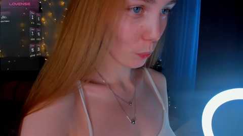 Media: Video of a young, fair-skinned woman with long, straight, reddish-blonde hair, wearing a white spaghetti-strap top, sitting indoors with a blue backdrop and a glowing ring light.