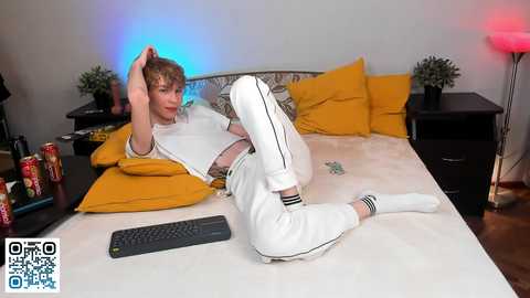 Media: Video of a young man with light skin and brown hair, lounging on a bed in a modern room with yellow pillows, blue and red lighting, and a black keyboard.