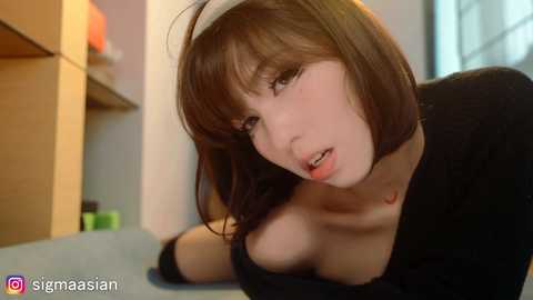 A video of a young, pale-skinned woman with short brown hair and a black choker, leaning forward seductively. She wears a black top exposing cleavage. The background shows a wooden shelf with orange objects.