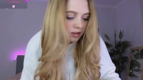 A video of a young woman with long, straight, blonde hair, wearing a white top, leaning forward, in a dimly lit room with a potted plant in the background.