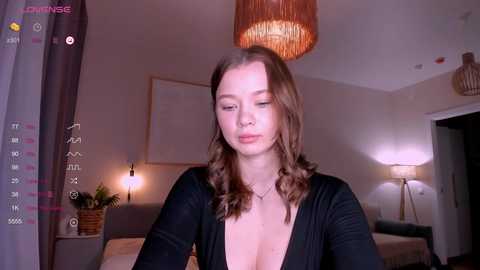 Media: A video of a young woman with wavy brown hair, wearing a low-cut black top, in a dimly lit living room with a modern decor.