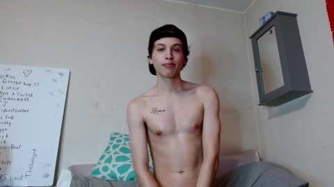 Media: Video of a shirtless, young, fit male with short dark hair, wearing a necklace, standing in a bedroom with white walls, a green patterned pillow, and a gray blanket.