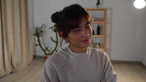 Media: Video of a young woman with medium skin tone, dark hair styled in a messy bun, wearing a cream-colored sweater, in a minimalist, warmly lit room with light wooden floors, beige curtains, and a potted plant.