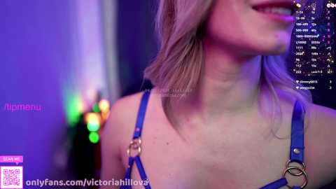 Media: Video of a smiling woman with fair skin, shoulder-length blonde hair, wearing a blue harness top. Background features colorful neon lights and a chat window with user names and messages.