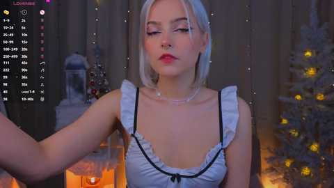 Media: Video of a fair-skinned, platinum blonde woman with medium-sized breasts, wearing a frilly white dress, standing in a dimly-lit room with lit candles, a Christmas tree, and a fireplace.