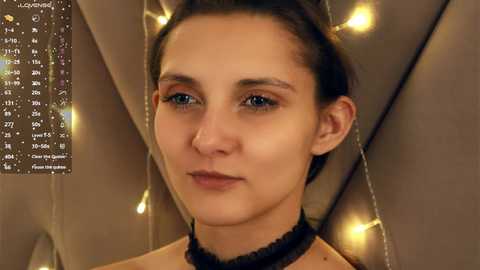 Video of a young woman with fair skin, brown hair, and blue eyes, wearing a black choker, standing in a dimly lit room with string lights in the background.