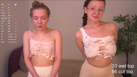 Media: Video of two young women, one with light skin, the other with medium skin, wearing torn, white crop tops and beige pants, standing indoors.