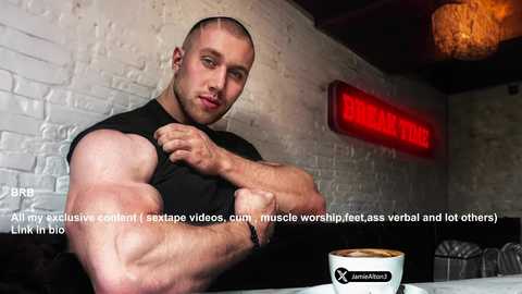 Video of a muscular man with a bald head and short beard, flexing his bicep in a black sleeveless shirt, sitting in a dimly lit cafe with a cup of coffee.