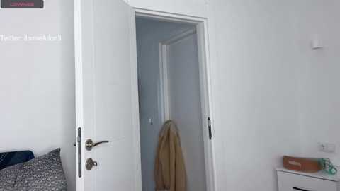 A video of a minimalist bedroom with a white door open, revealing a blonde woman in a beige robe. The room has white walls and a small bedside table with a book.