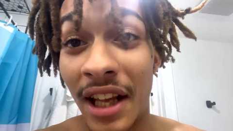 Media: Video of a young Black man with light brown skin and short dreadlocks, smiling widely, in a bathroom with a blue shower curtain and white towels.