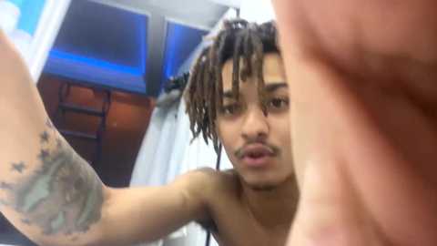 Media: Video of a topless Black man with dreadlocks and a tattoo on his forearm, holding a phone to take a selfie. Background shows a modern, minimalist room with blue lighting and a white curtain.