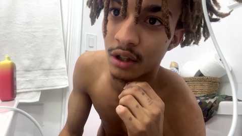 Media: Video of a shirtless young Black man with short dreadlocks, light brown skin, and a mustache, holding his chin in a bathroom with a white towel, soap dispenser, and wicker basket.