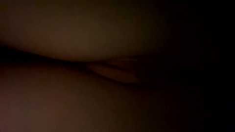 A dimly lit, grainy video of a naked, light-skinned person's buttocks and upper thighs. The skin appears smooth and the background is dark and indistinct, with a slight reddish hue.