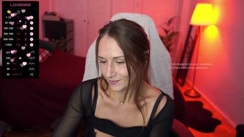 Media: Video of a fair-skinned woman with long brown hair, wearing a sheer black top, seated on a bed with a white blanket, bathed in warm red light.