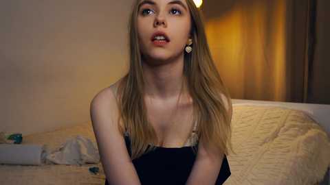 Media: Video of a young blonde woman with fair skin, wearing a black tank top, sitting on a beige quilted bed in a dimly lit room.